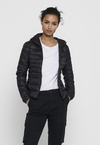 Black puffer jacket with a hood, featuring a quilted design, front zipper, and elastic cuffs. Worn over a white t-shirt and black pants.