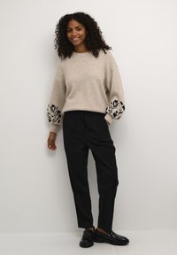 Pull beige avec broderies florales noires sur les manches, associé à un pantalon large noir et des chaussures vernies noires. Tissu texturé, coupe décontractée.