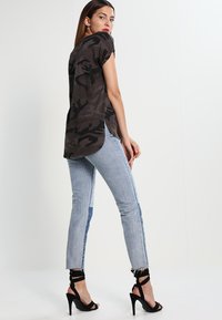 Mønsteret i kamuflasje i mørkegrått, kortermet, avrundet kant. Kombinert med lyseblå høytlivsjeans med kontrasterende sidepaneler og opprevet kant.
