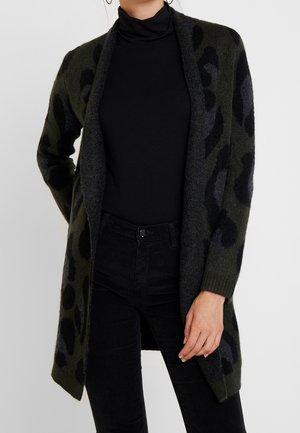 Woman wearing dark green and black patterned open-front cardigan over black turtleneck and black pants, standing with one hand at side.