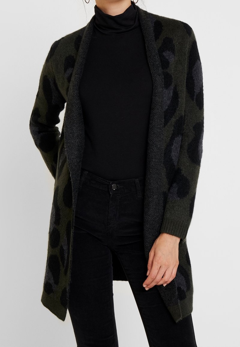 Femme portant un cardigan ouvert à motifs vert foncé et noir par-dessus un col roulé noir et un pantalon noir, debout avec une main le long du corps.