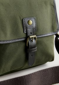 Olivengrüne Stofftasche mit einem schwarzen Lederträger, ausgestattet mit einer Messingschnalle und einem Druckknopfverschluss, sichtbare Nähte und einer strukturierten Oberfläche.