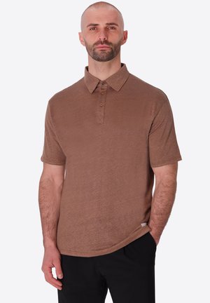 Uomo con la barba, indossa una polo marrone a maniche corte e pantaloni neri, in piedi con una mano in tasca su uno sfondo semplice.