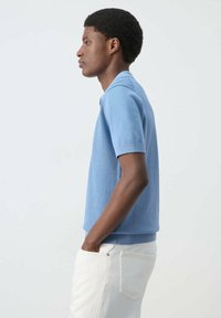 Joven con un suéter de manga corta texturizado azul claro y pantalones blancos, de pie de perfil con las manos en los bolsillos y un fondo liso.