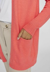 Korallfarbener Strickcardigan mit Rippbündchen und einer Vordertasche, kombiniert mit crèmefarbenen Hosen und einem weißen Hemd. Weicher, strukturierter Stoff.