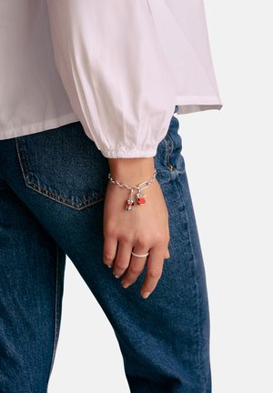 Silbernes Charm-Armband mit bunten Anhängern, die rote Akzente setzen. Eine Hand, die das Armband trägt, ruht auf Jeansstoff, ein silberner Ring ist sichtbar.