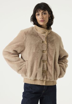 Vrouw draagt een beige namaakbontjack met drukknopen en elastische tailleband, gecombineerd met donkerblauwe spijkerbroek, staand met handen in de zakken.