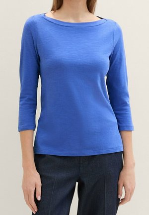 Kobieta ubrana w dopasowaną, niebieską bluzkę z długim rękawem i dekoltem w kształcie łodzi oraz ciemnoniebieskie spodnie, stojąca na tle jasnego, jednolitego tła.