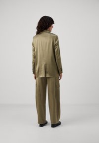 Completo satinato verde oliva con giacca sartoriale e pantaloni cargo larghi. La giacca ha una sola fessura; i pantaloni presentano tasche laterali e una vestibilità rilassata.