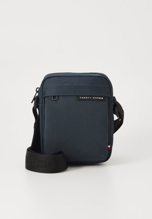 Navy blue crossbody bag made of textured fabric, featuring a zipper, adjustable strap, and a logo tag along the front with red and white accents.