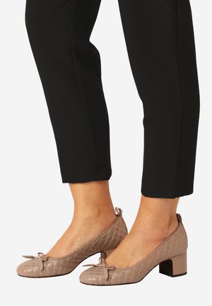 Gesteppte beige Schuhe mit niedrigem Absatz und einer Schleife oben, getragen zu schwarzen, kurz geschnittenen Hosen, gezeigt von den Knöcheln abwärts.