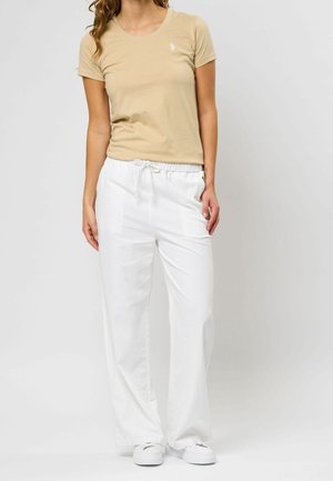 Femme portant un t-shirt beige à manches courtes, un pantalon blanc avec cordon de serrage et des baskets blanches, debout devant un fond clair uni.