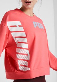 Korálová mikina s bílým a modrým logem "PUMA", má uvolněný střih, dlouhé rukávy, žebrované manžety a měkkou texturu látky.