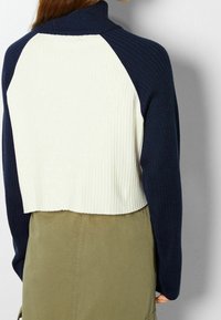 Croptröja med marinblå och krämfärgade ribbade texturer, hög hals och raglanärmar. Matchas med en olivgrön underdel.