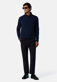 Maglione con collo a zip blu navy realizzato in tessuto knit, abbinato a pantaloni scuri e sneaker nere. Il modello indossa occhiali da sole, in piedi su uno sfondo neutro.