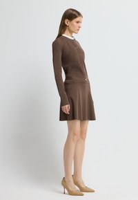 Bruine gebreide jurk met lange mouwen, ronde halslijn, knopen aan de voorkant en getailleerde taille. Geaccentueerd met een riem met een metalen gesp. Draagt met beige hakken.