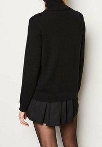 Pull en tricot noir à col roulé avec manches longues, présentant une silhouette ajustée. Associé à une mini jupe plissée noire et des collants noirs.