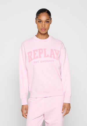 Frau trägt hellrosa Sweatshirt und Hose mit dem Text „REPLAY NOT ORDINARY“ auf dem Sweatshirt, steht vor einem schlichten hellen Hintergrund.