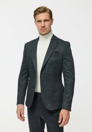 Man wearing dark gray suit with black pocket square and light beige turtleneck, standing against a plain light background.