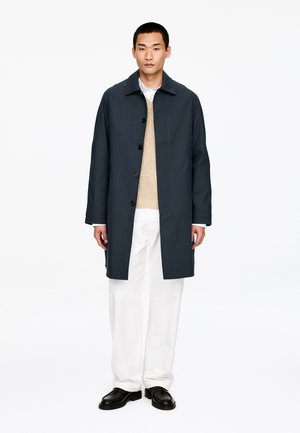 Mann trägt einen dunkelblauen, geknöpften Mantel über einem beigen Pullover, weiße Hose und schwarze Schuhe, steht vor einem schlichten weißen Hintergrund.