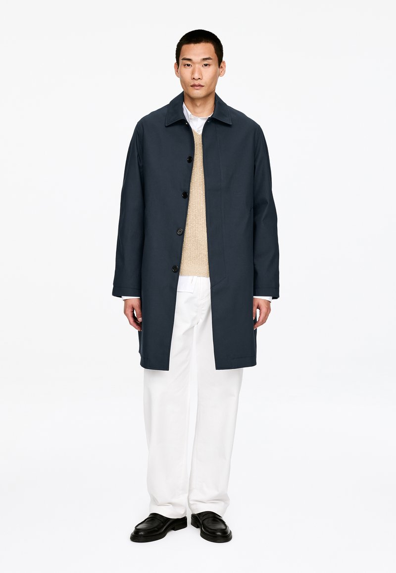 Mand iført en mørk marineblå knapjakke over en beige sweater, hvide bukser og sorte sko, stående mod en ensfarvet hvid baggrund.