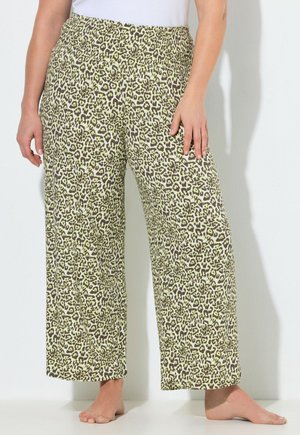 Vrouw die losse broek met luipaardprint en een elastische tailleband draagt, staat blootsvoets tegen een effen witte muur.