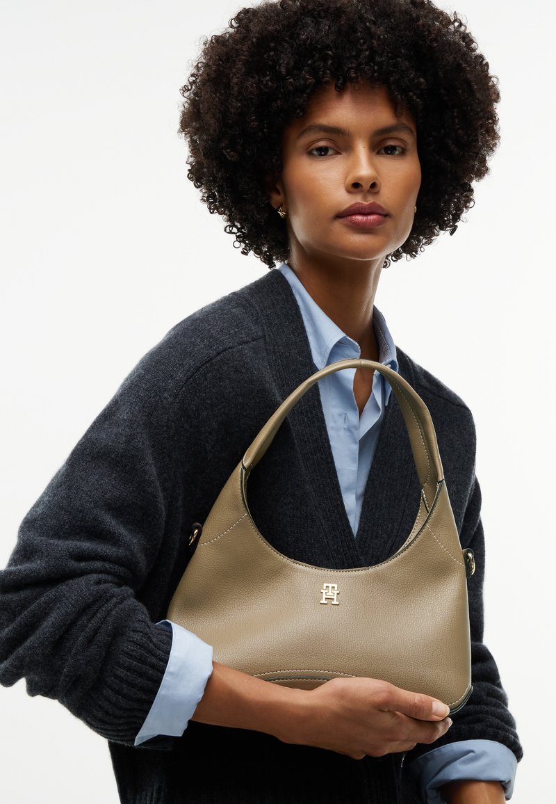 Femme aux cheveux bouclés tient un sac à main en cuir marron clair avec un logo doré, portant un cardigan gris foncé sur une chemise bleu clair.