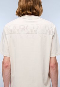 Beige T-skjorte med korte ermer og en teksturert grafisk trykk på ryggen som viser ordet "NAPAPIJRI" med en subtil logo under.