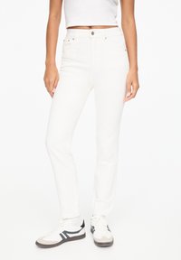 Vaqueros de mezclilla blanca con un corte clásico, que cuentan con cierre de botón, cinco bolsillos y costuras mínimas. Llevados con zapatillas casuales.