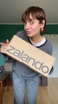 Kartonasta škatla Zalando drži se ob strani, z velikimi sivečimi črkami. Maneken nosi dolgi rokav s potiskom v mornarsko modri in beli barvi ter svetlo sivo hlače.
