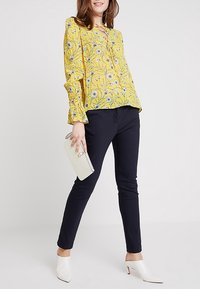 Blouse jaune à motifs floraux avec des manches à volants, associée à un pantalon slim noir, un sac à main blanc et des mules blanches à bout pointu.