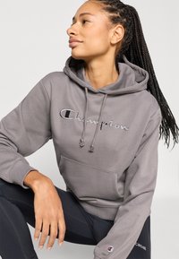 Γκρι φούτερ με κουκούλα και τσέπη καγκουρό, με κεντημένο το λογότυπο "Champion". Διαθέτει κουκούλα με κορδόνι και υφασμάτινη υφή. Ορατά μαύρα λεγινγκ.