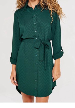 Vrouw die een donkergroene, lange-mouwen, jurk met knopen draagt, met een ceintuur in de taille en opgerolde mouwen, tegen een effen achtergrond.