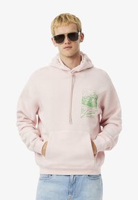 Sudadera rosa claro con un bolsillo frontal, capucha con cordón y un estampado gráfico verde en el pecho. Material suave y corte relajado.