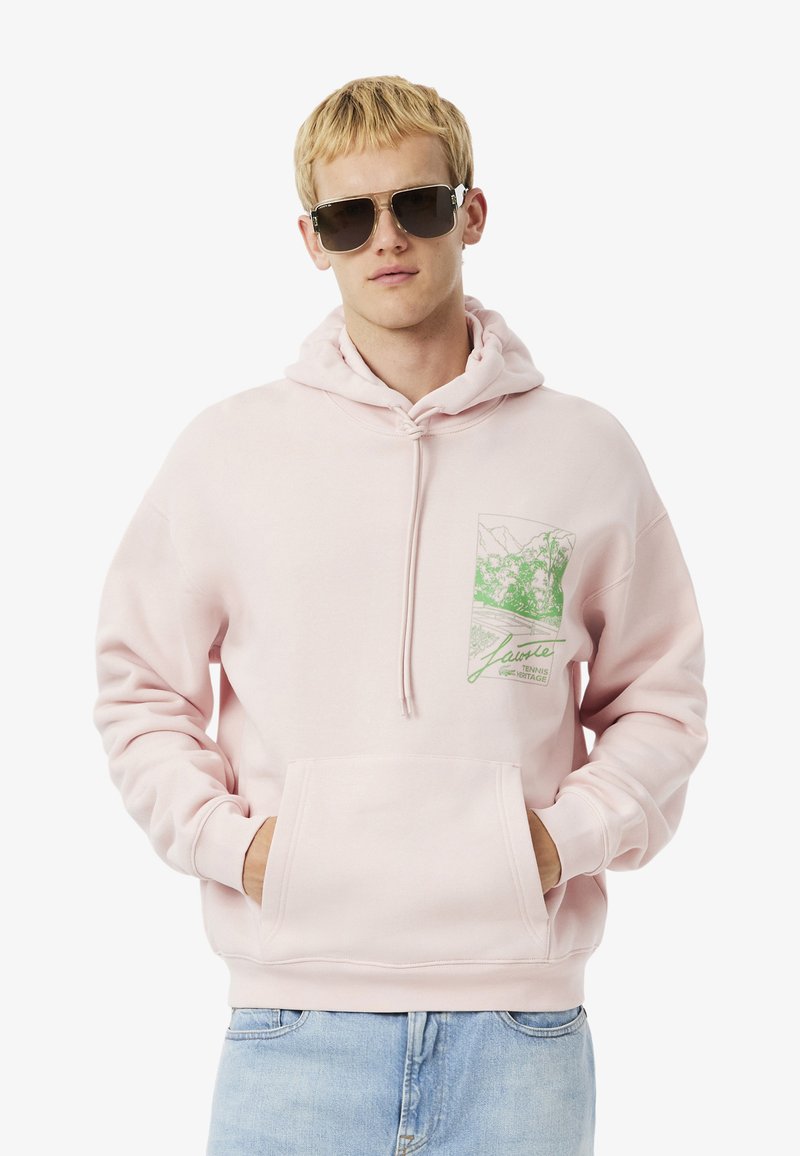 Sudadera rosa claro con un bolsillo frontal, capucha con cordón y un estampado gráfico verde en el pecho. Material suave y corte relajado.