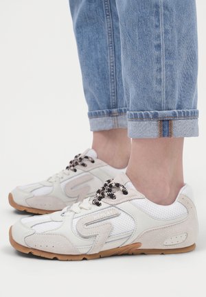 Baskets blanches et beiges avec des lacets noirs et blancs portées avec un jean bleu retroussé sur un fond blanc uni.