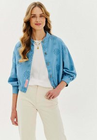 Světlemodrá bombera s květinovou výšivkou, patentkami a žebrovanými manžetami, nošená přes bílé tričko a světle béžové kalhoty.