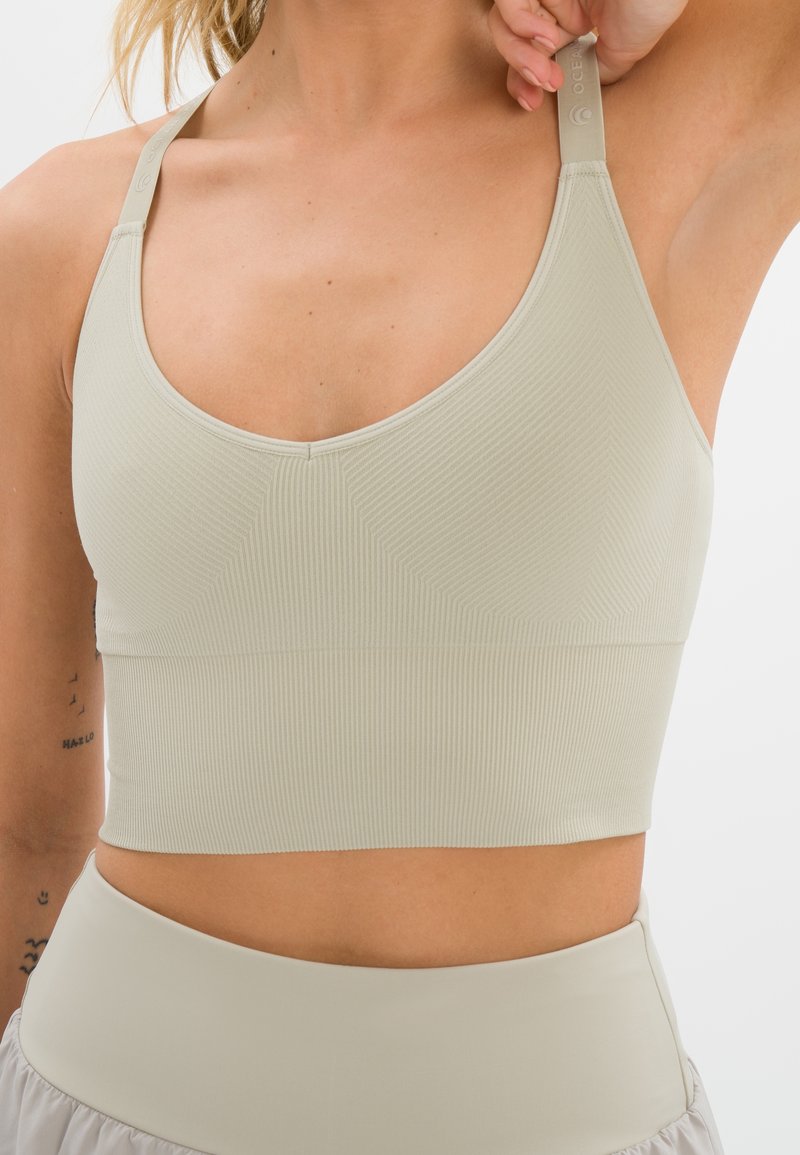 Femme portant un crop top beige clair côtelé avec de larges bretelles élastiques, associé à une jupe ou un pantalon taille haute, bras levé à hauteur d'épaule.