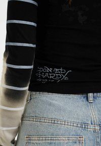 Maglietta nera a maniche lunghe con strisce grigie, caratterizzata da un gradiente sfumato, e un logo bianco sul lato sinistro in basso, abbinata a jeans in denim chiaro.