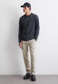 Donkergrijze shirt met lange mouwen en een klein logo, beige broek en zwarte schoenen. Het model draagt een bijpassende donkere pet en staat tegen een neutrale achtergrond.