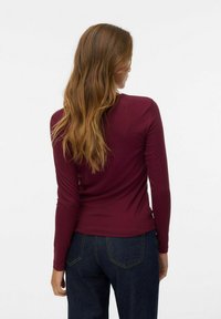 Femme avec de longs cheveux ondulés portant un haut ajusté bordeaux à manches longues et un jean bleu foncé, tournant le dos contre un fond blanc uni.