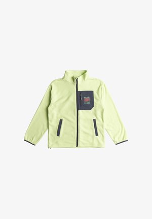 Jasnozielona polarowa kurtka z wysokim kołnierzem, zamek błyskawiczny i kieszeń na piersi z granatowym panelem oraz kolorową grafiką. Noszona z niebieskimi dżinsami.