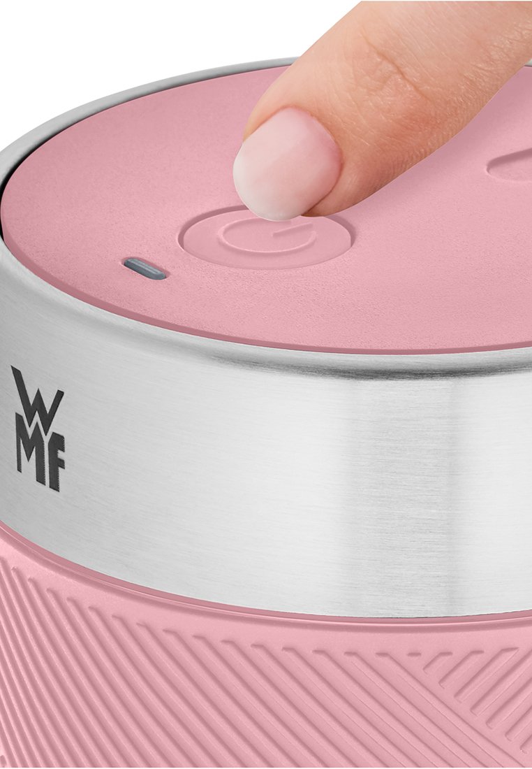 Ein Finger drückt den Power-Button eines pinkfarbenen, strukturierten tragbaren WMF-Geräts mit einem Edelstahlband und einer kleinen Anzeigeleuchte.