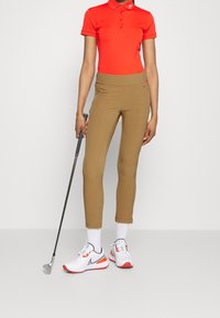 Camisa polo roja con tres botones, pantalones de golf ajustados marrones, calcetines blancos hasta el tobillo y zapatos deportivos grises con detalles naranjas, sosteniendo un palo de golf.