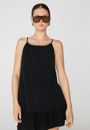 Donna che indossa occhiali da sole scuri oversize, orecchini a cerchio in argento e un vestito nero senza maniche con spalline sottili su sfondo semplice.