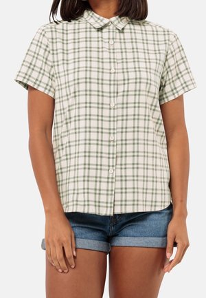 Mujer con una camisa de manga corta abotonada con un patrón de cuadros verdes y blancos y pantalones cortos de mezclilla azul remangados.