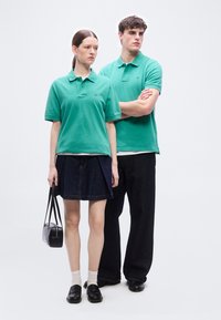 Νέα γυναίκα και άνδρας που φορούν τιρκουάζ πόλο πουκάμισα· η γυναίκα κρατάει μαύρη τσάντα, φοράει σκουρόχρωμη φούστα και μοκασίνια, ο άνδρας φοράει σκουρόχρωμο φαρδύ παντελόνι και παπούτσια.