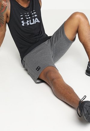 Maglietta sportiva nera senza maniche con logo "UA", abbinata a pantaloncini grigi con una piccola etichetta del marchio nera, indossata con scarpe da ginnastica nere.