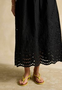 Melnā izšūtā kokvilnas maksi kleita ar izgrieztiem ziedu rakstiem apakšā, kombinēta ar dzeltenām čībiņām ar mezglu detaļām.