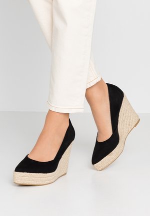 Jambes portant un pantalon blanc et des talons compensés noirs avec des semelles plateformes en corde tressée sur un sol gris.